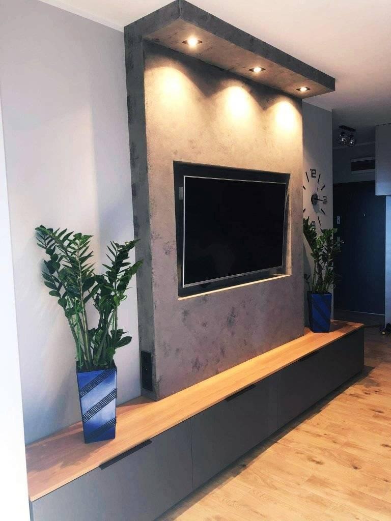 Nowoczesny salon z szarą ścianą TV, oświetleniem punktowym i drewnianą podłogą, uzupełniony roślinami w niebieskich donicach z geometrycznym wzorem.