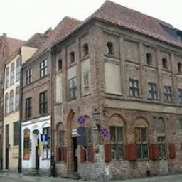 Kamienica z cegły z charakterystycznymi łukami nad oknami i czerwonymi okiennicami, widoczna tabliczka z nazwą ulicy i zakaz parkowania.