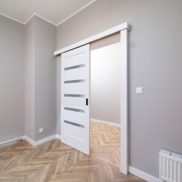 mieszkaniepodklucz.com Paweł Styga - Białe, przesuwne drzwi z poziomymi przeszkleniami, częściowo otwarte, w szarym pokoju z parkietem ułożonym we wzór jodełki. Widoczny grzejnik i listwy przypodłogowe.