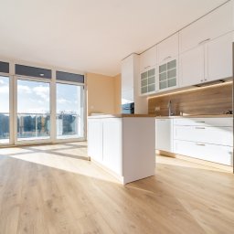 mieszkaniepodklucz.com Paweł Styga - Jasna, przestronna kuchnia z białymi szafkami, wyspą kuchenną i parkietem, oświetlona naturalnym światłem wpadającym przez duże okna balkonowe.