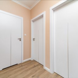 mieszkaniepodklucz.com Paweł Styga - Trzy białe, minimalistyczne drzwi wewnętrzne z metalowymi klamkami w korytarzu o jasnych ścianach i podłodze imitującej drewno.