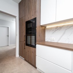 mieszkaniepodklucz.com Paweł Styga - Nowoczesna kuchnia z białymi szafkami i blatem z drewna, zabudowany piekarnik i mikrofalówka w brązowej obudowie, marmurowe płytki na ścianie, widok na korytarz z otwartych drzwi.