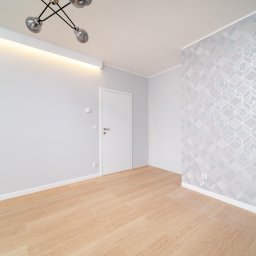mieszkaniepodklucz.com Paweł Styga - Jasne wnętrze po odświeżeniu: parkiet w odcieniach beżu, ściany pomalowane na szaro z geometrycznym wzorem tapety, białe drzwi i nowoczesna lampa sufitowa.