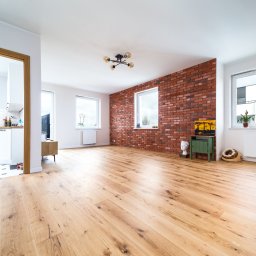 mieszkaniepodklucz.com Paweł Styga - Jasny salon z drewnianą podłogą, ścianą z czerwonej cegły, widokiem na kuchnię przez otwarte drzwi i regałem z drewnianymi półkami