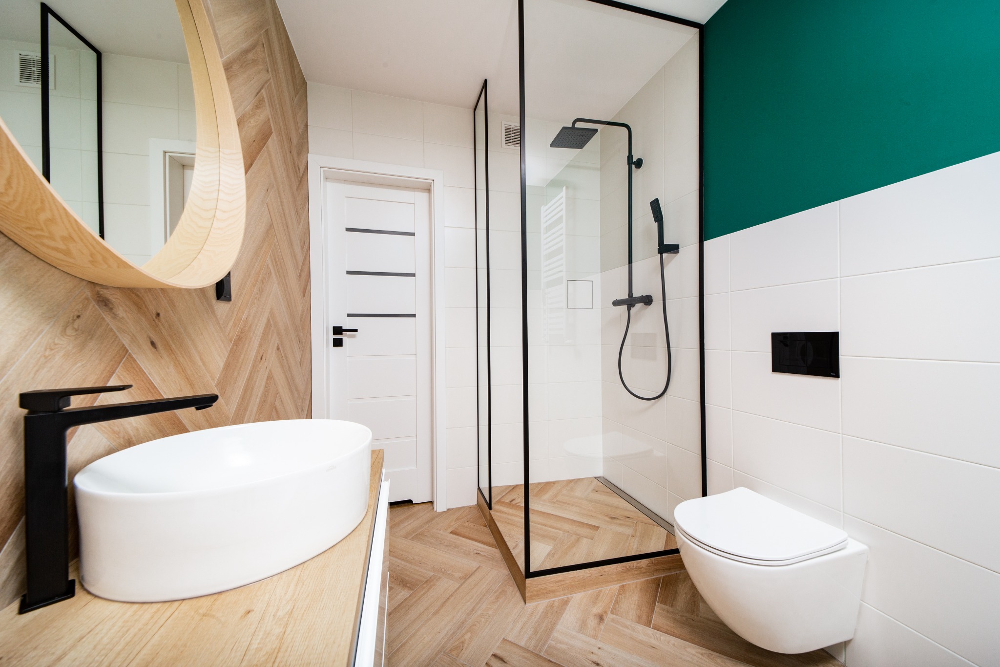 Nowoczesna łazienka z drewnianą podłogą ułożoną w jodełkę, okrągłym lustrem z drewnianą ramą, kabiną prysznicową z czarnymi profilami i białą ceramiką.