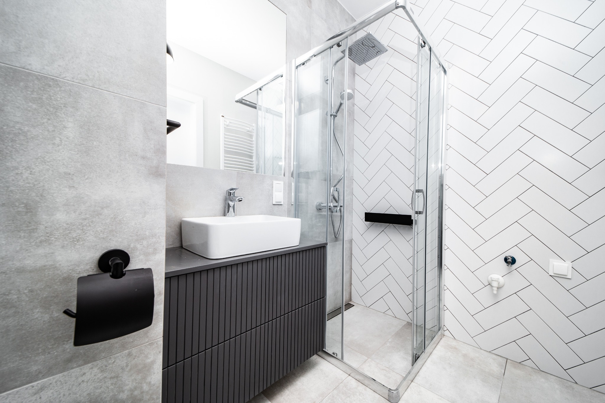 Nowoczesna łazienka z białą umywalką nablatową, szarą szafką z pionowymi żłobieniami, kabiną prysznicową w rogu oraz białymi płytkami ułożonymi w jodełkę na ścianie prysznica, uzupełniona o czarny...