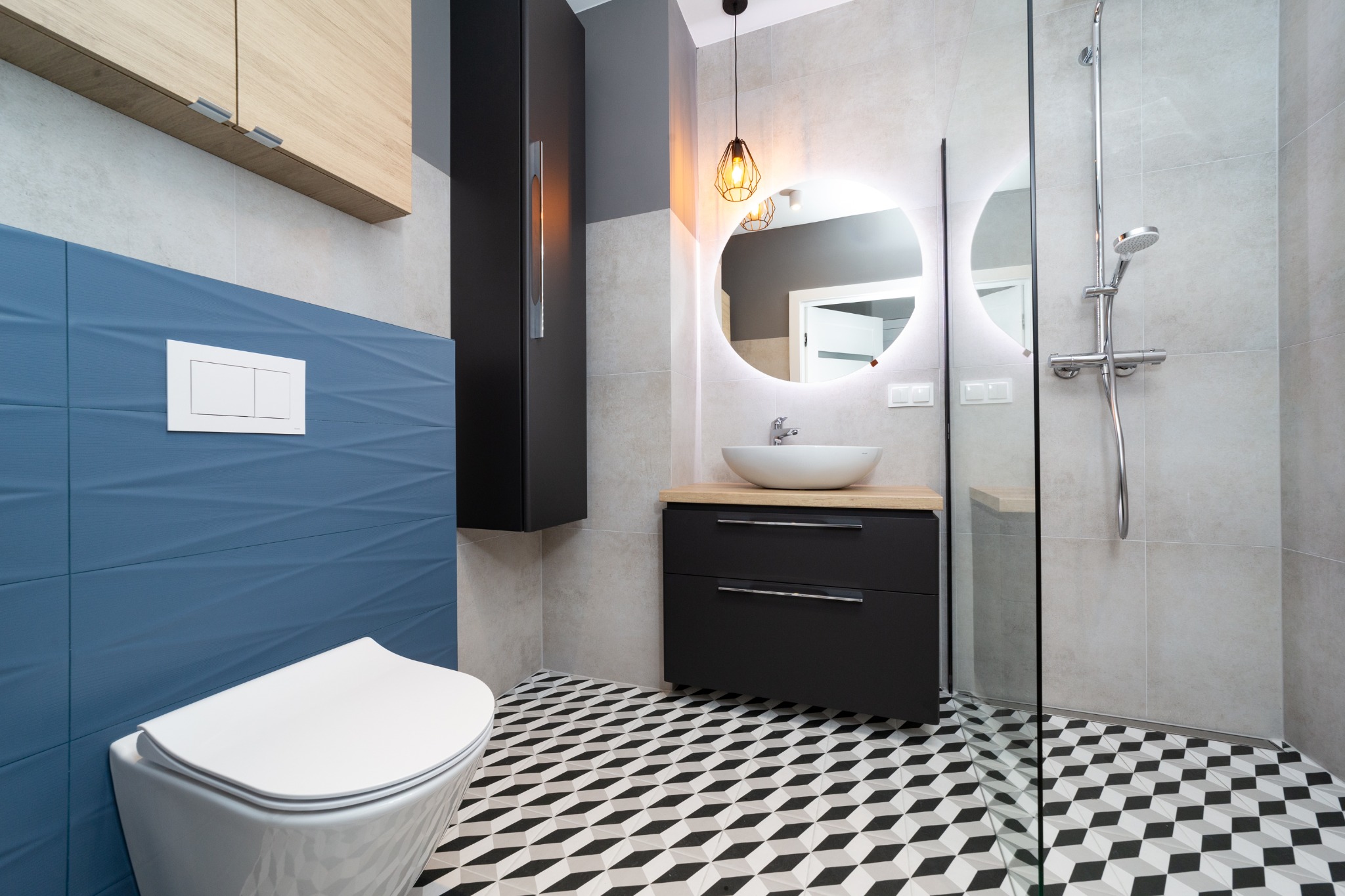 Nowoczesna łazienka z niebieską ścianą z geometrycznym wzorem, wiszącą toaletą, szafką z umywalką na drewnianym blacie, okrągłym lustrem z podświetleniem LED i podłogą w geometryczny wzór 3D.