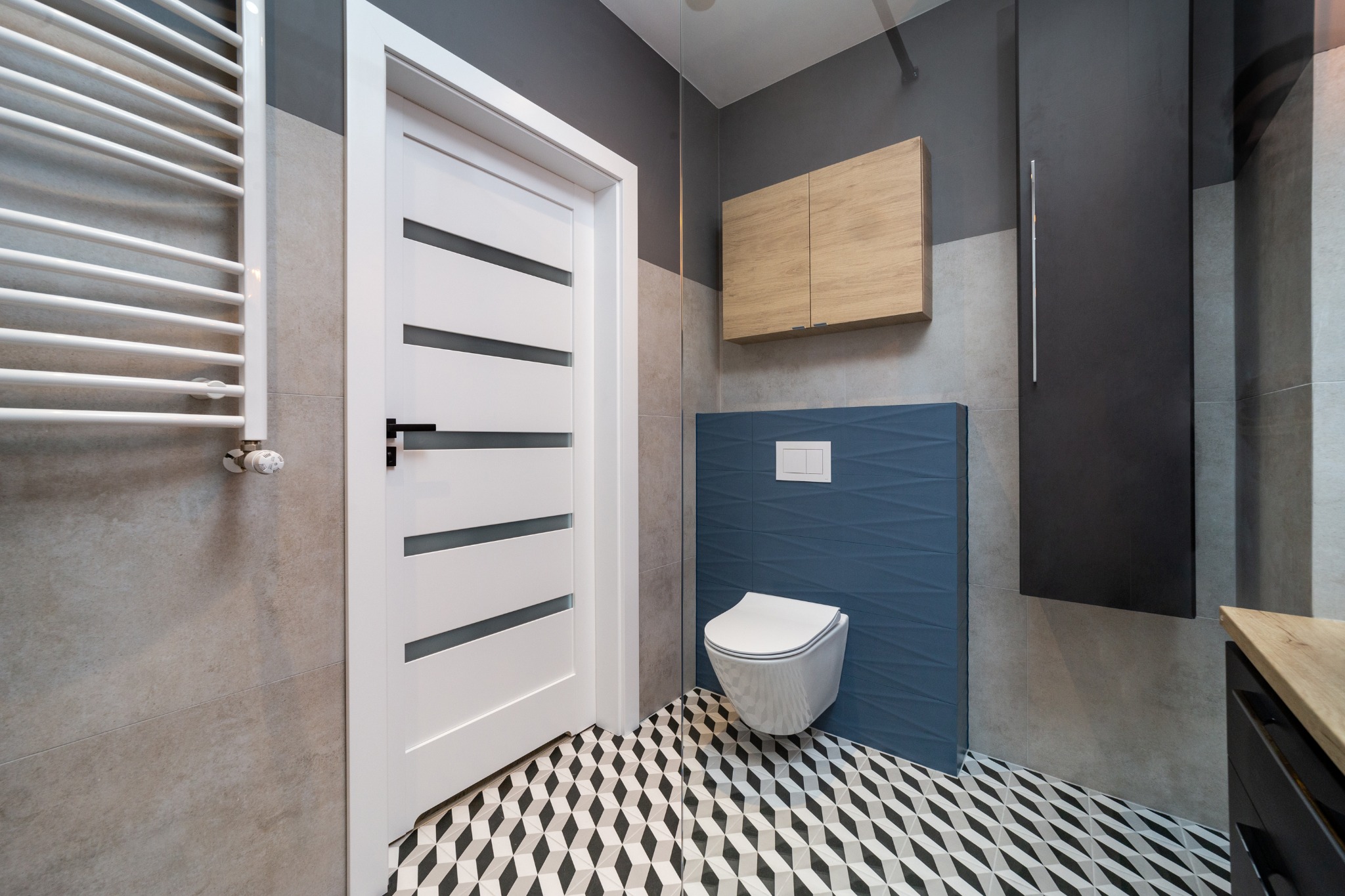 Nowoczesna łazienka z białą podwieszaną toaletą na tle niebieskich płytek 3D, białe drzwi z poziomymi przeszkleniami i grzejnik drabinkowy.