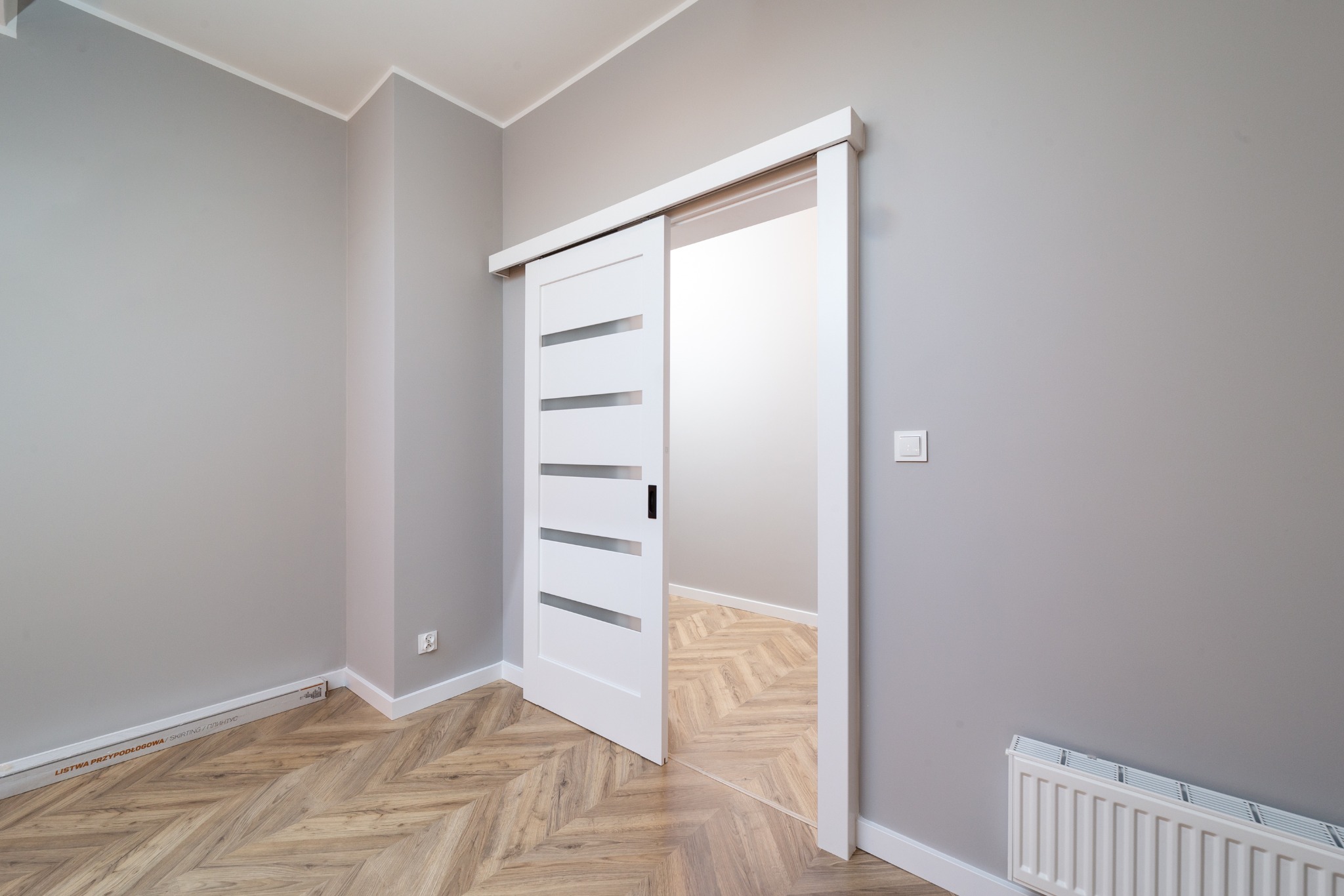 Białe, przesuwne drzwi z poziomymi przeszkleniami, częściowo otwarte, w szarym pokoju z parkietem ułożonym we wzór jodełki. Widoczny grzejnik i listwy przypodłogowe.
