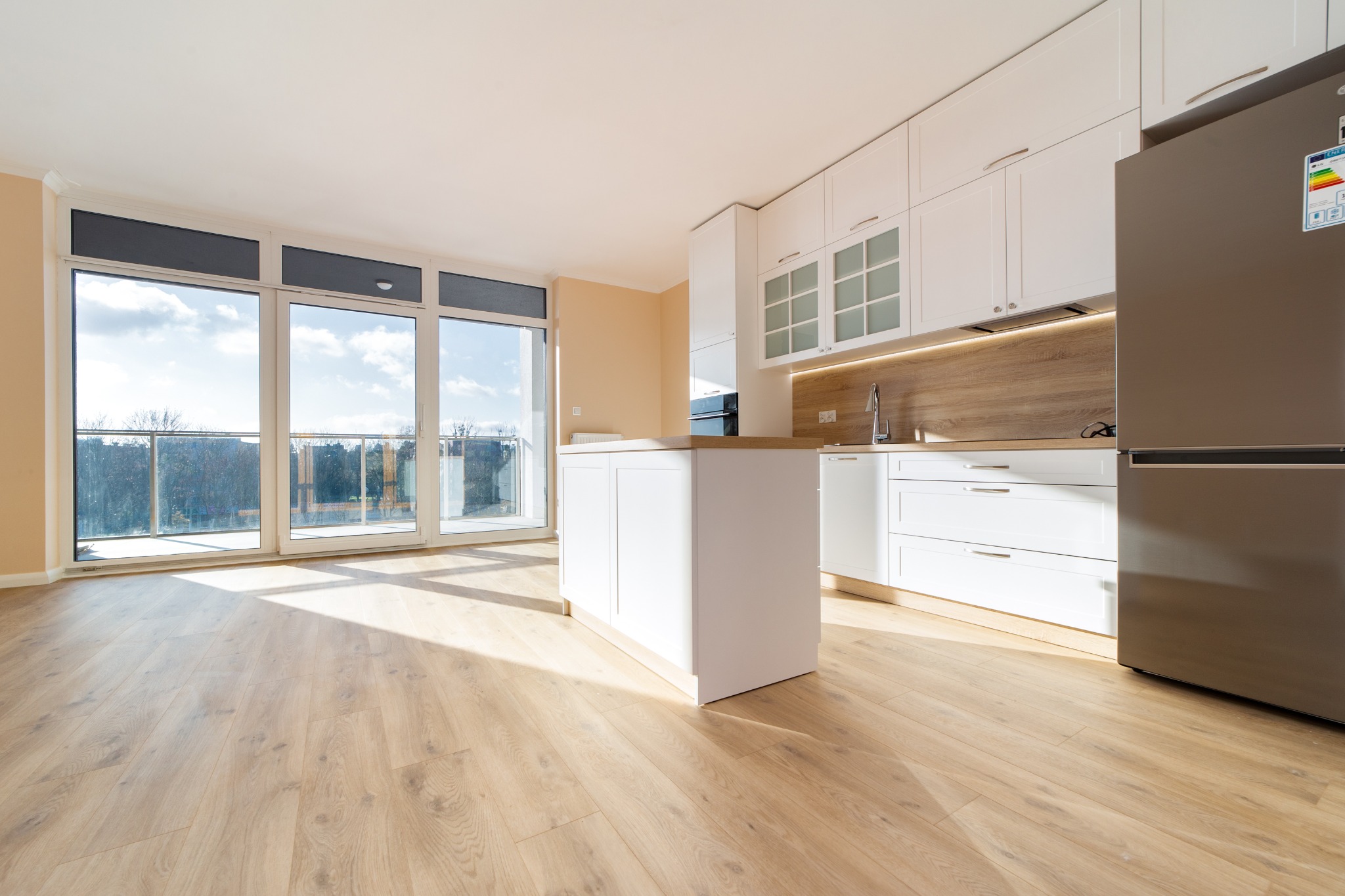 Jasna, przestronna kuchnia z białymi szafkami, wyspą kuchenną i parkietem, oświetlona naturalnym światłem wpadającym przez duże okna balkonowe.