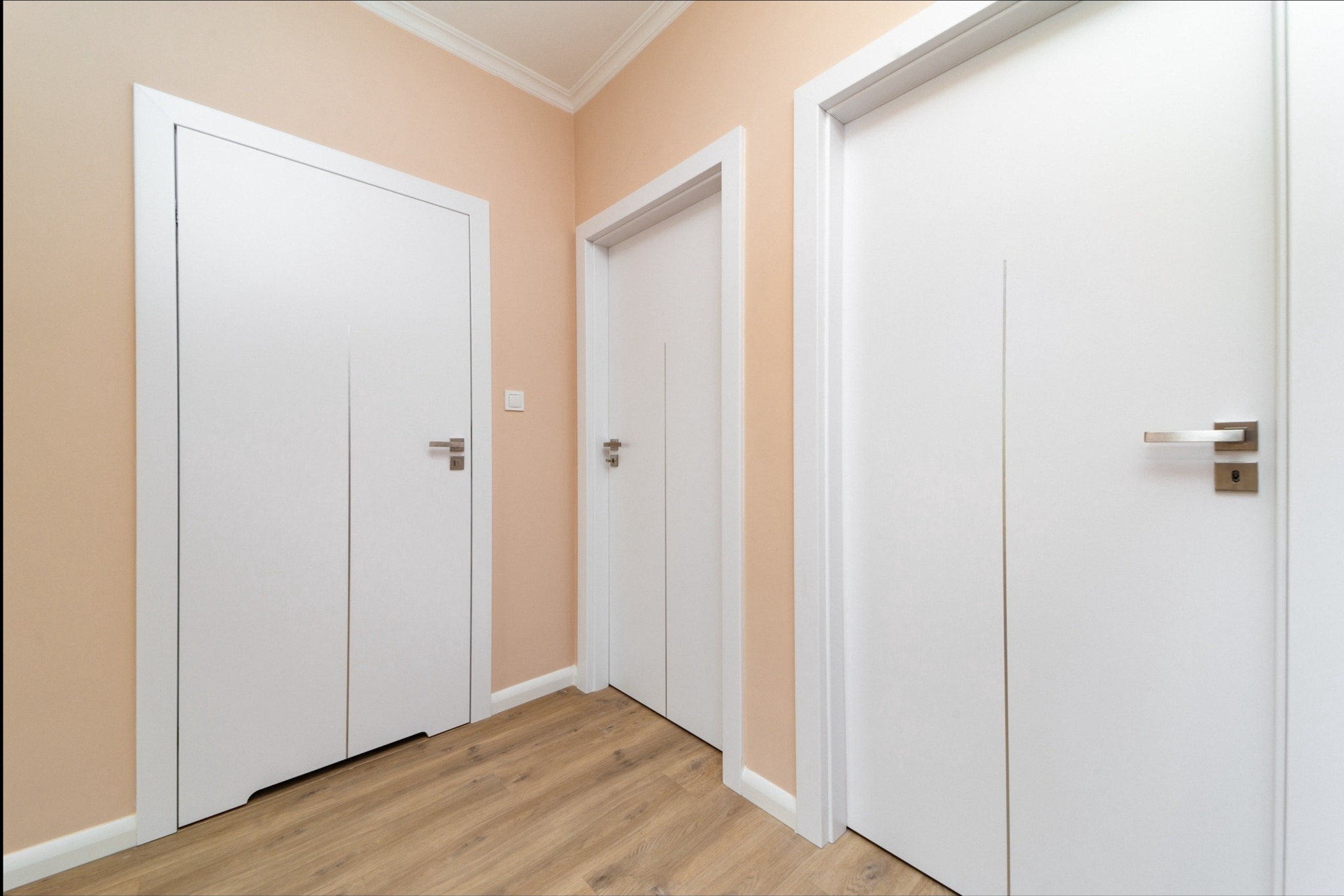 Trzy białe, minimalistyczne drzwi wewnętrzne z metalowymi klamkami w korytarzu o jasnych ścianach i podłodze imitującej drewno.