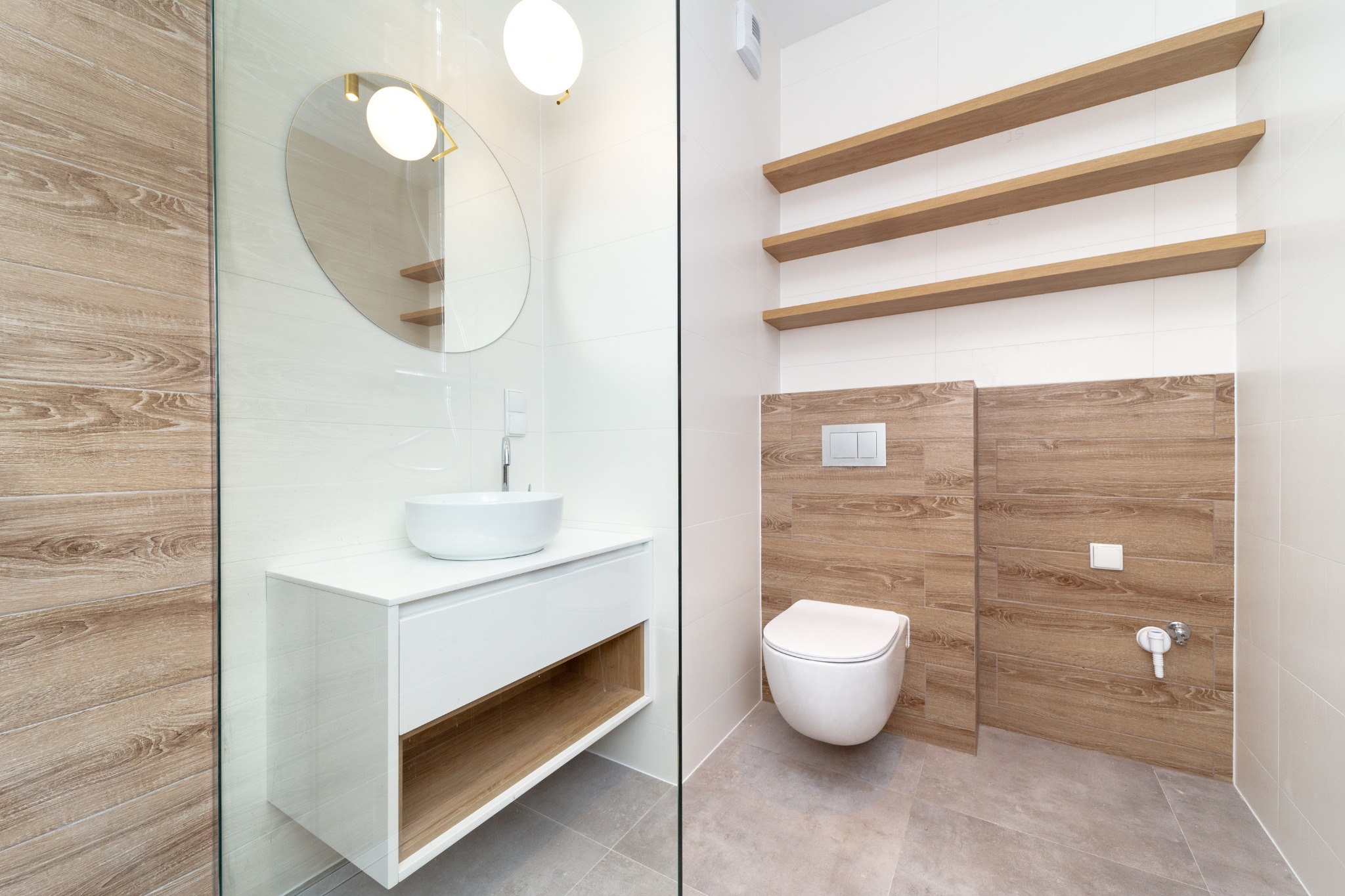 Nowoczesna łazienka z białymi płytkami, drewnopodobnymi panelami, okrągłym lustrem z podświetleniem, wiszącą miską WC i drewnianymi półkami.