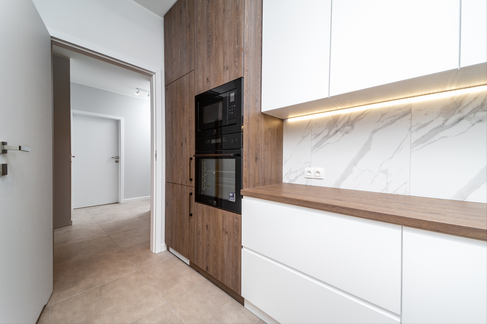 Nowoczesna kuchnia z białymi szafkami i blatem z drewna, zabudowany piekarnik i mikrofalówka w brązowej obudowie, marmurowe płytki na ścianie, widok na korytarz z otwartych drzwi.