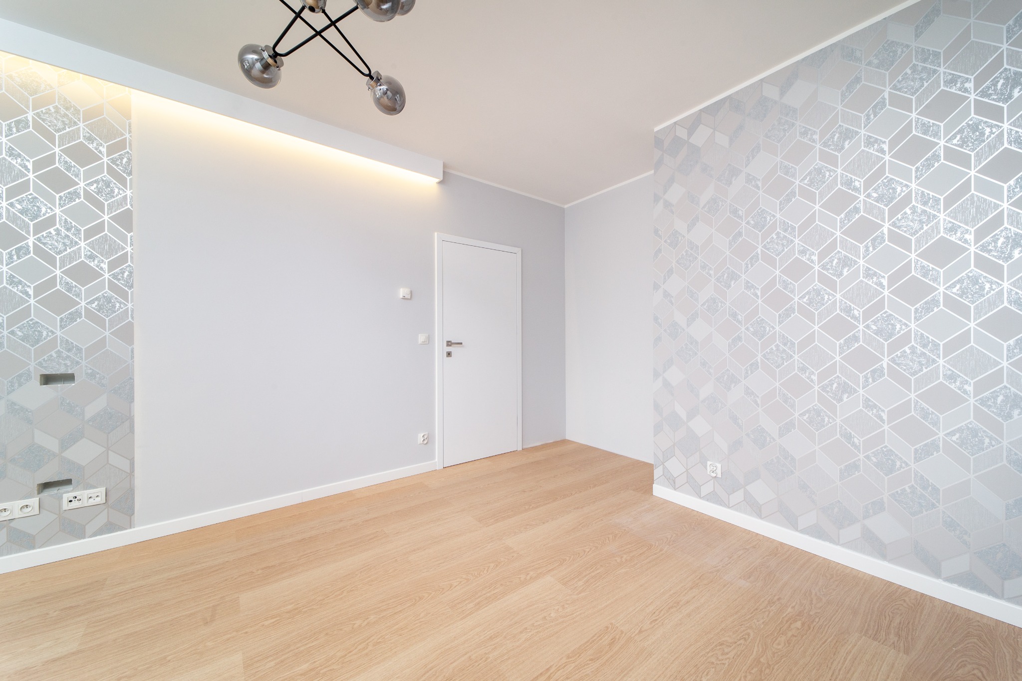 Jasne wnętrze po odświeżeniu: parkiet w odcieniach beżu, ściany pomalowane na szaro z geometrycznym wzorem tapety, białe drzwi i nowoczesna lampa sufitowa.