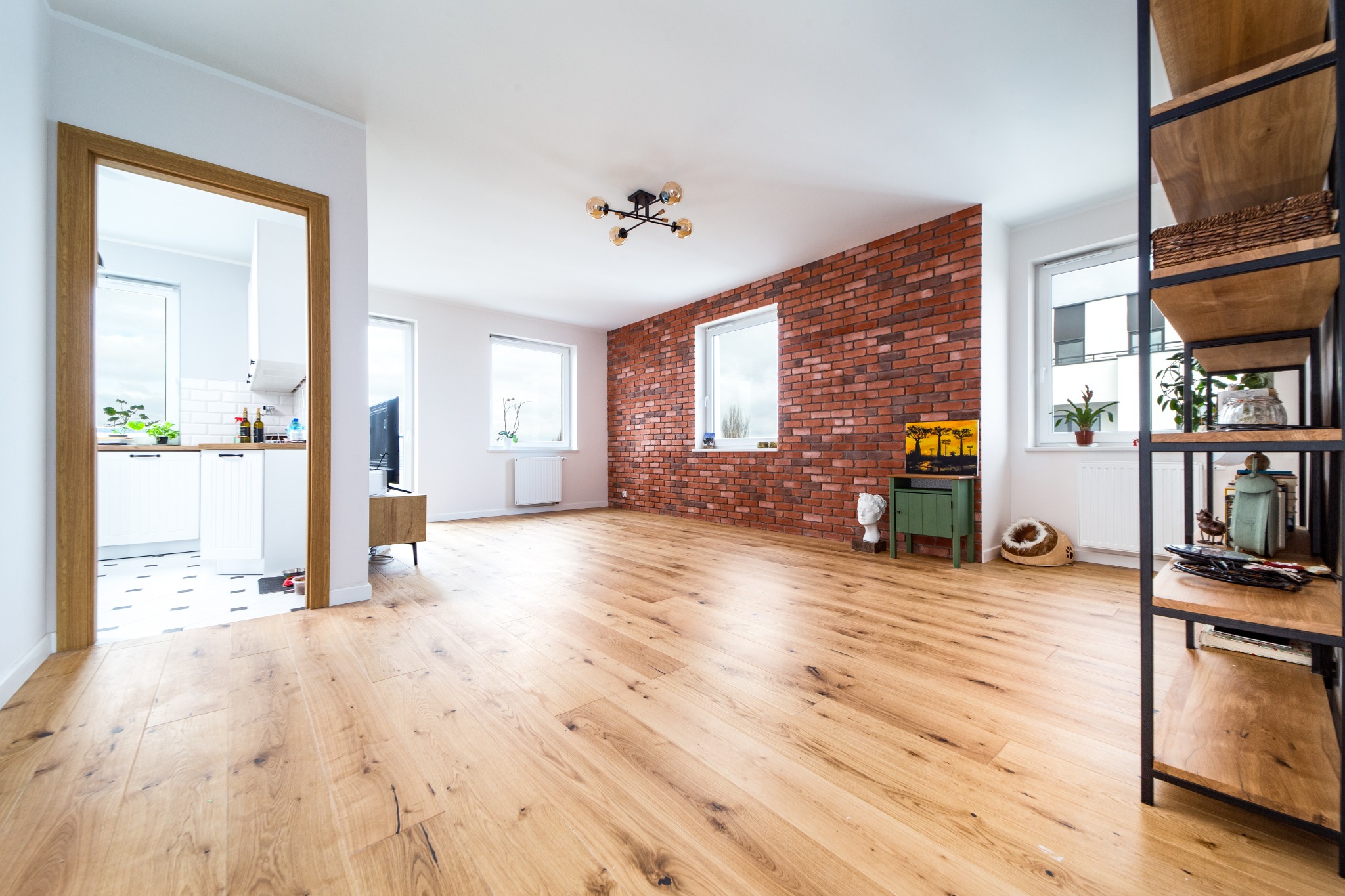 Jasny salon z drewnianą podłogą, ścianą z czerwonej cegły, widokiem na kuchnię przez otwarte drzwi i regałem z drewnianymi półkami
