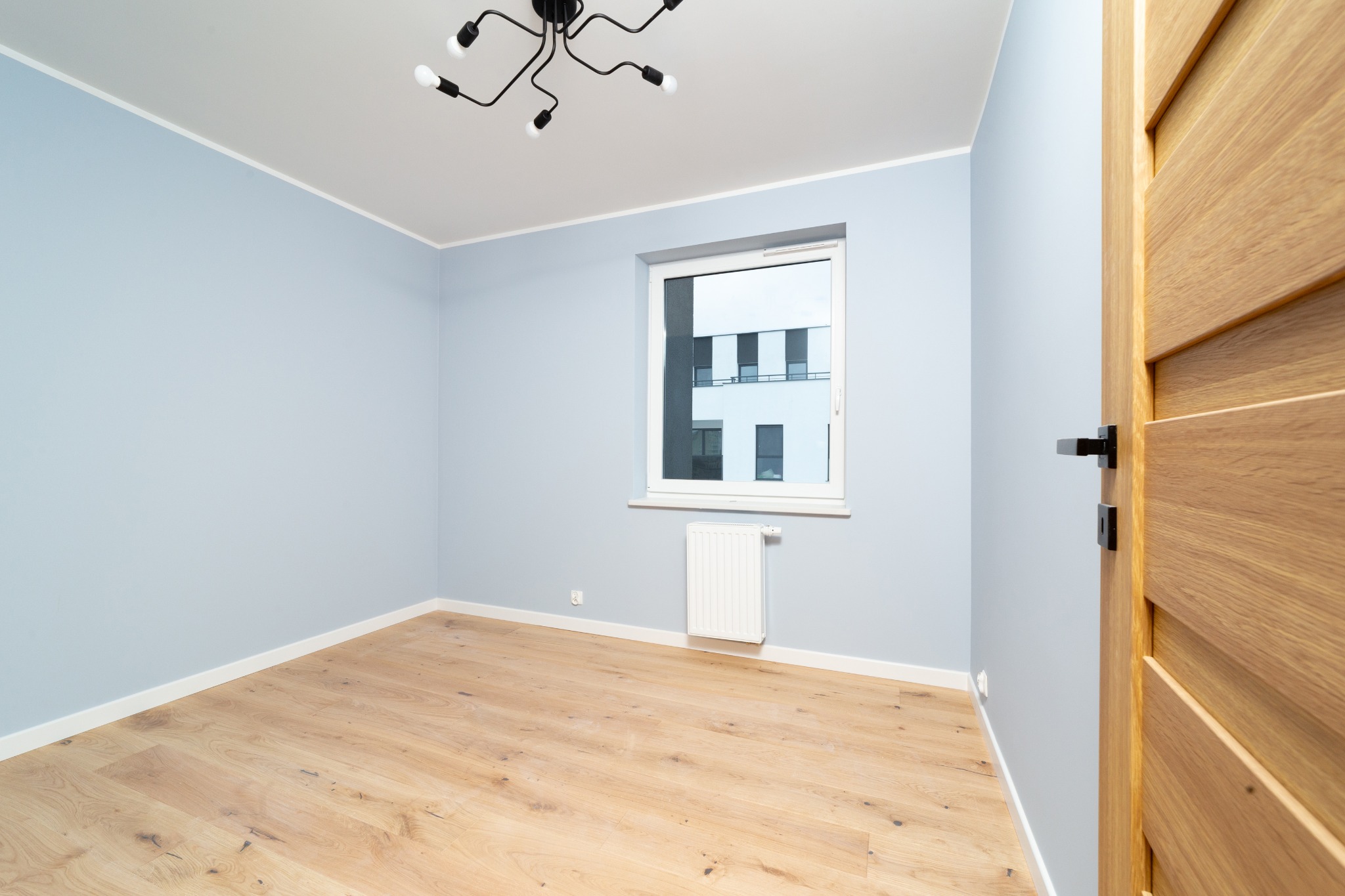 Pusty pokój po odświeżeniu: świeżo pomalowane błękitne ściany, jasny parkiet, białe listwy przypodłogowe, nowoczesna lampa sufitowa i dębowa, wewnętrzna stolarka drzwiowa z czarną klamką.