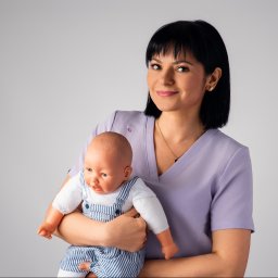 Logopeda, Neurologopeda Paulina Łataś - Uśmiechnięta kobieta w fioletowym uniformie trzyma lalkę niemowlęcą w niebiesko-białym kombinezonie na jasnym tle. Złoty łańcuszek i kolczyki dodają blasku.