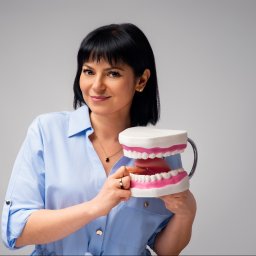 Logopeda, Neurologopeda Paulina Łataś - Uśmiechnięta kobieta w błękitnej koszuli prezentuje model szczęki z różowymi dziąsłami na jasnym tle. Widoczny złoty pierścionek i naszyjnik.