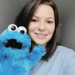 Uśmiechnięta kobieta z brązowymi włosami trzyma niebieską pacynkę z dużymi oczami, prawdopodobnie podczas sesji terapeutycznej lub zabawy.