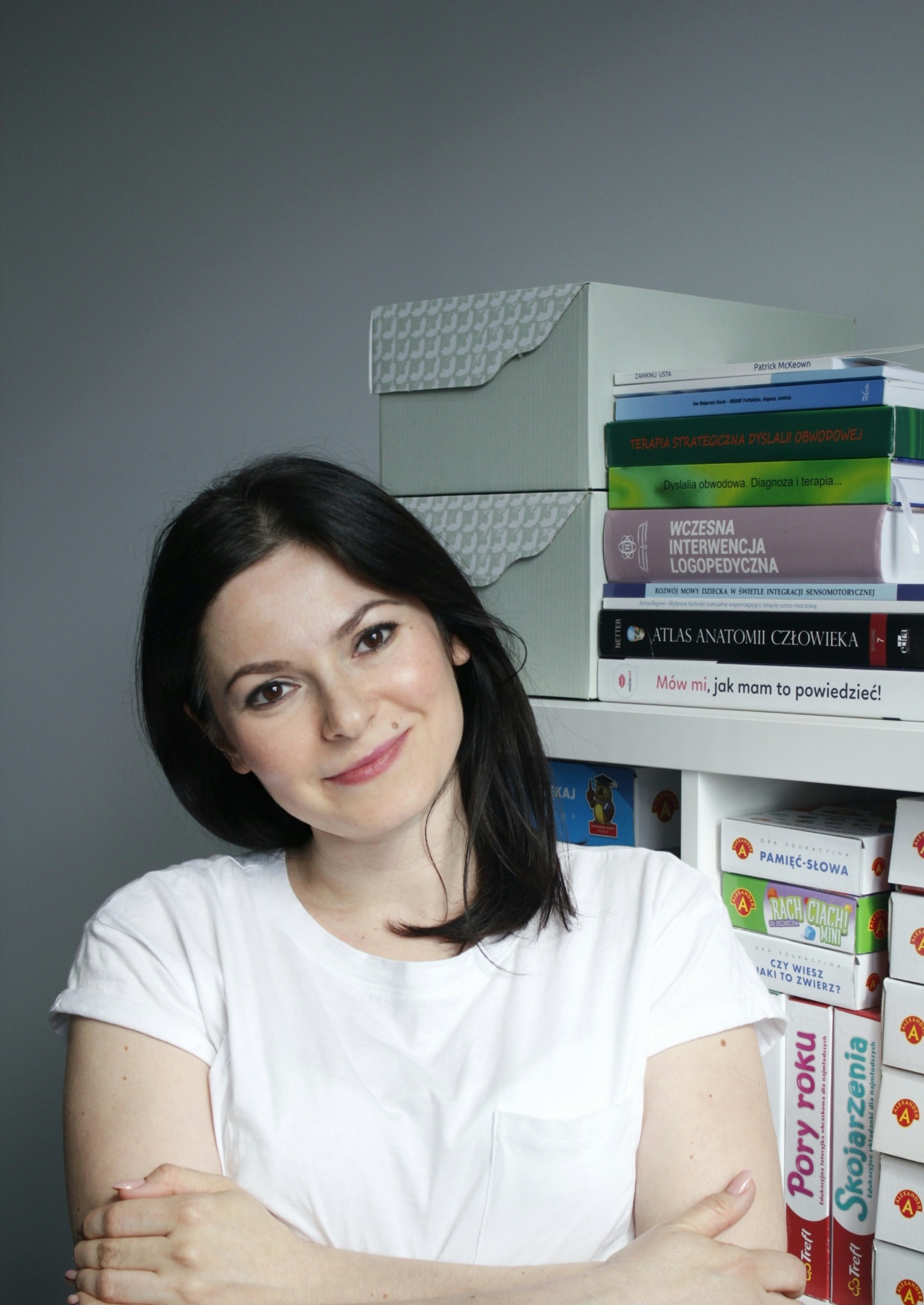 Uśmiechnięta logopedka w białej koszulce, z książkami i grami logopedycznymi w tle, w tym 'Atlas Anatomii Człowieka', 'Wczesna interwencja logopedyczna' oraz gry edukacyjne 'Pamięć-Słowa'...