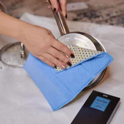 Dłoń kobiety w trakcie czyszczenia dna stalowego garnka niebieską ściereczką z żółtymi kropkami; obok pudełko z napisem 'Splitter'.