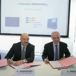 Dwóch mężczyzn w garniturach podpisuje dokumenty na tle ekranu z logotypami instytucji finansowych w jasnym pomieszczeniu.