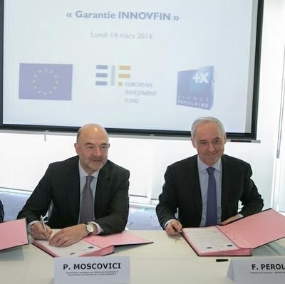 Dwóch mężczyzn w garniturach podpisuje dokumenty na tle ekranu z logotypami instytucji finansowych w jasnym pomieszczeniu.