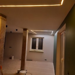 Usługi remontowo budowlane - Wnętrze w trakcie remontu z widocznymi profilami LED na suficie, zabezpieczonymi folią słupami i puszkami z farbą na podłodze.