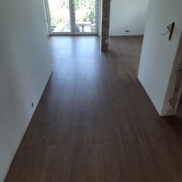 Usługi remontowo budowlane - Ułożona, brązowa podłoga imitująca drewno w jasnym pomieszczeniu w trakcie remontu; widoczny słup zabezpieczony folią, okno z widokiem na zieleń oraz wystające przewody elektryczne.