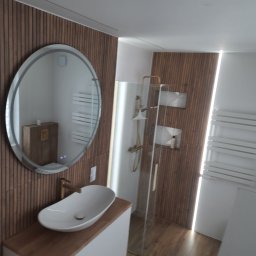 Usługi remontowo budowlane - Nowoczesna łazienka z owalną umywalką nablatową, okrągłym lustrem z podświetleniem LED i kabiną prysznicową ze złotymi elementami. Ściany wykończone drewnopodobnymi panelami.