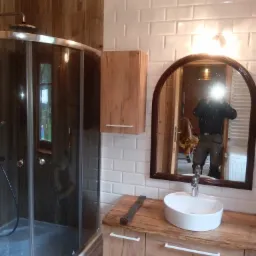 Nowoczesna łazienka z kabiną prysznicową z ciemnego szkła, umywalką nablatową na drewnianym blacie i białymi płytkami typu metro na ścianach. W lustrze odbicie osoby w roboczym stroju. Na blacie...