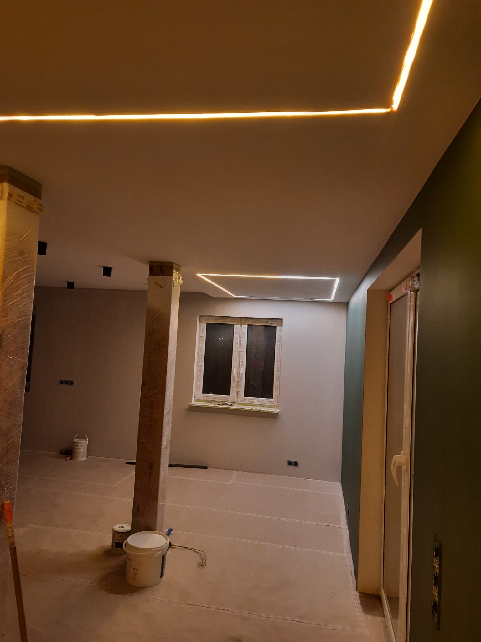 Wnętrze w trakcie remontu z widocznymi profilami LED na suficie, zabezpieczonymi folią słupami i puszkami z farbą na podłodze.