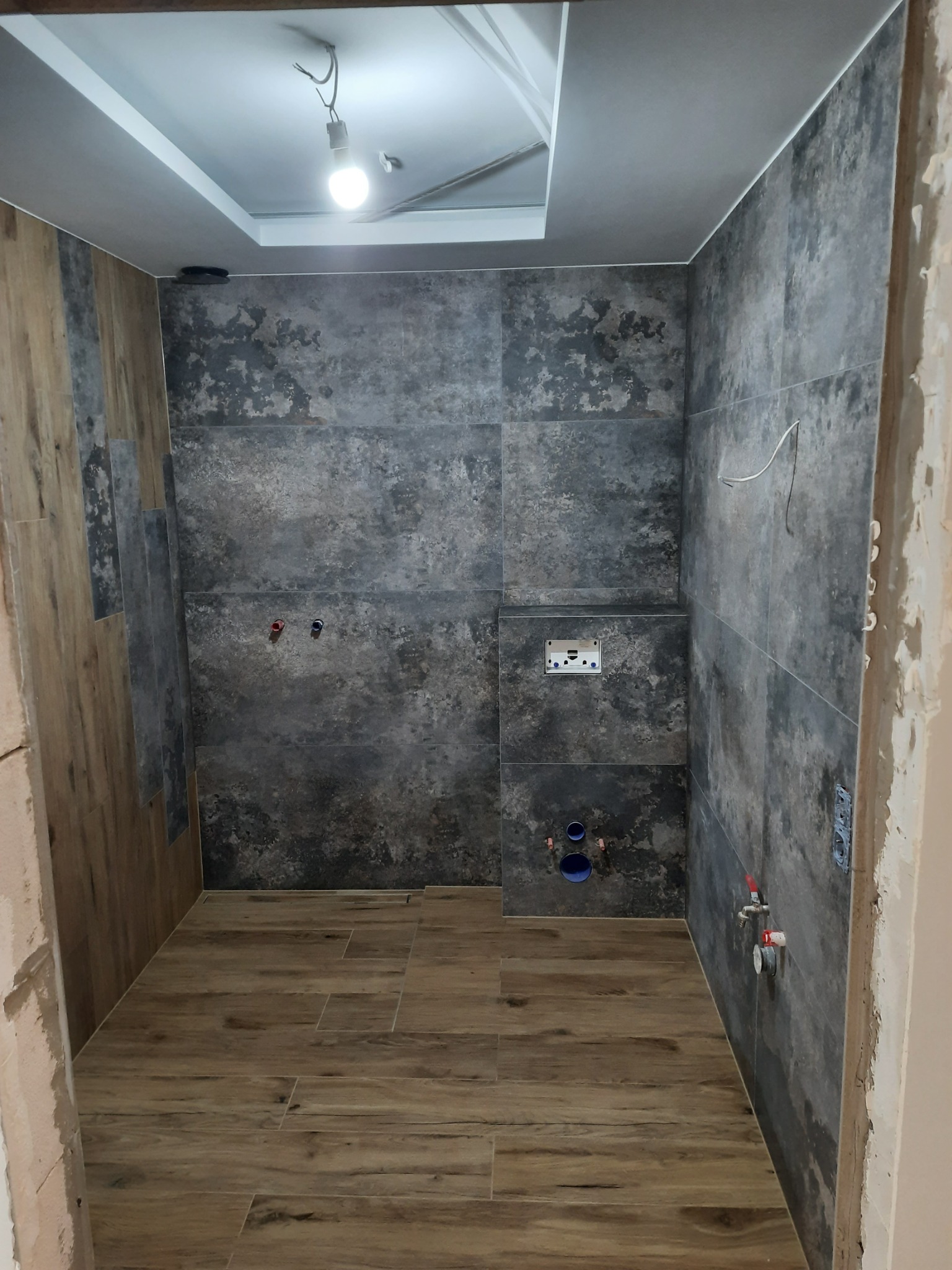 Wnętrze łazienki w trakcie remontu, widoczne szare płytki na ścianach z otworami na instalacje, drewnopodobna podłoga, surowy stan wykończenia przy framudze drzwi.