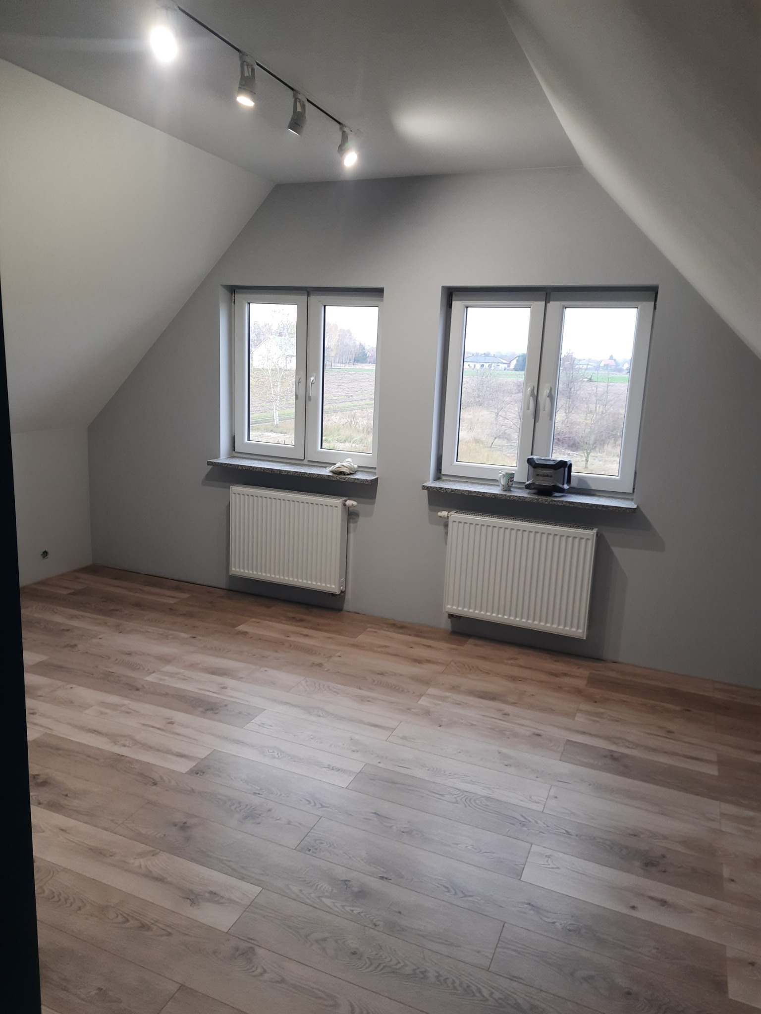 Pokój na poddaszu z drewnianą podłogą, szarą ścianą i dwoma oknami z widokiem na krajobraz wiejski, oświetlony reflektorami sufitowymi.