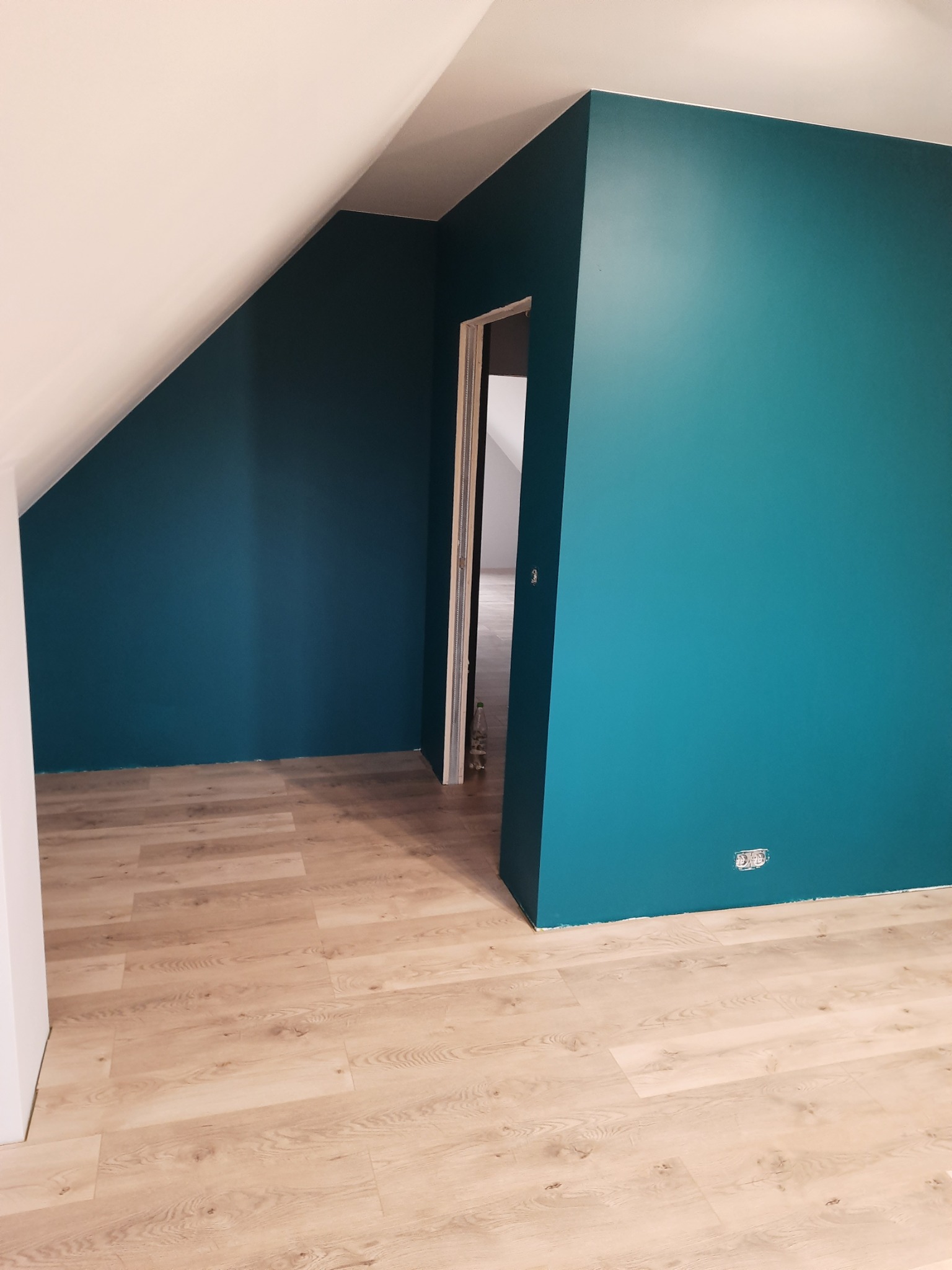 Pomalowane na turkusowo ściany w pokoju na poddaszu z jasną podłogą panelową i widocznym gniazdkiem elektrycznym.