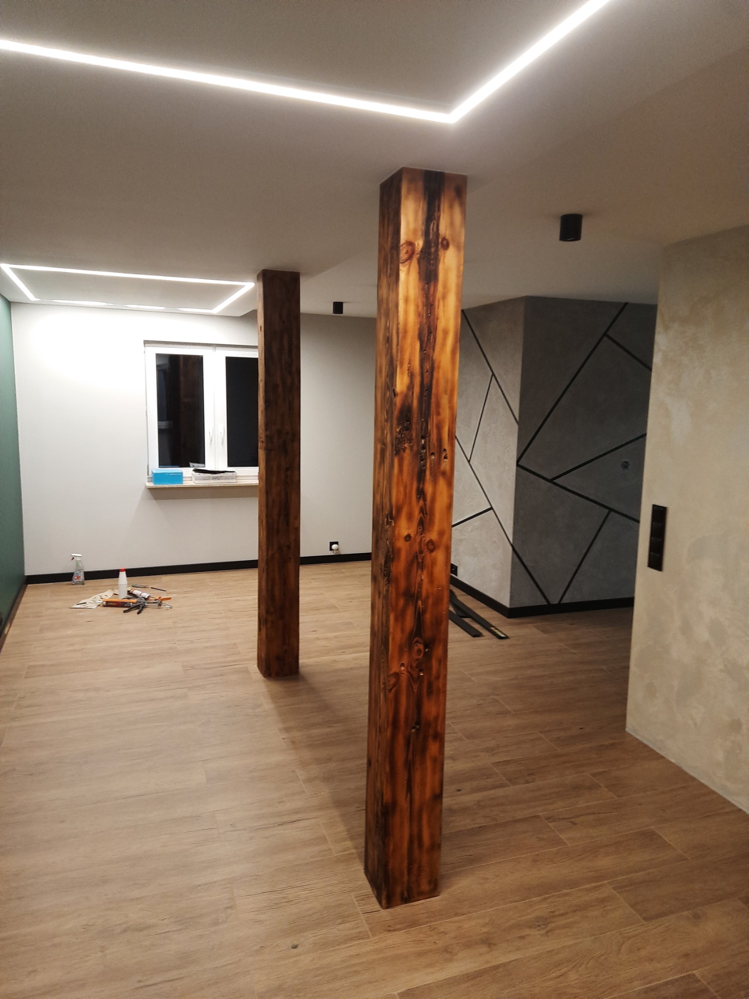 Wnętrze po remoncie z drewnopodobną podłogą, dwoma drewnianymi słupami dekoracyjnymi, oświetleniem LED w suficie i ścianą z geometrycznym wzorem.