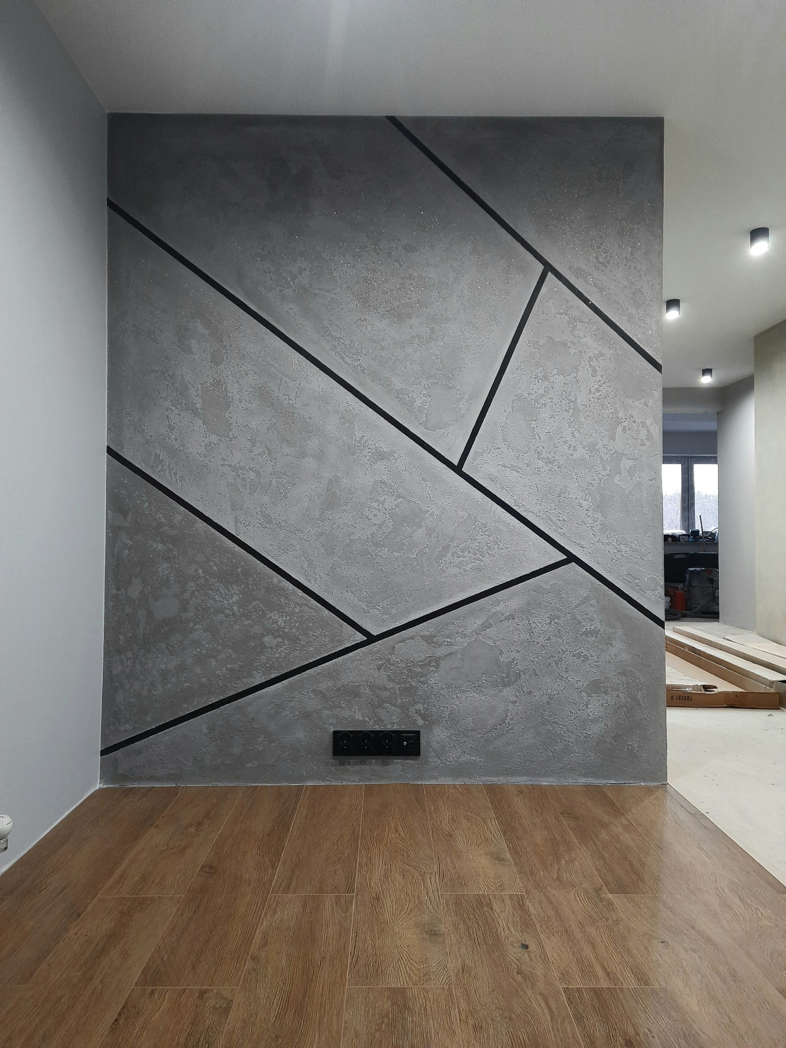 Ściana z dekoracyjną szarą strukturą i czarnymi, geometrycznymi liniami, brązowa podłoga imitująca drewno, w tle widoczny korytarz z oświetleniem punktowym.