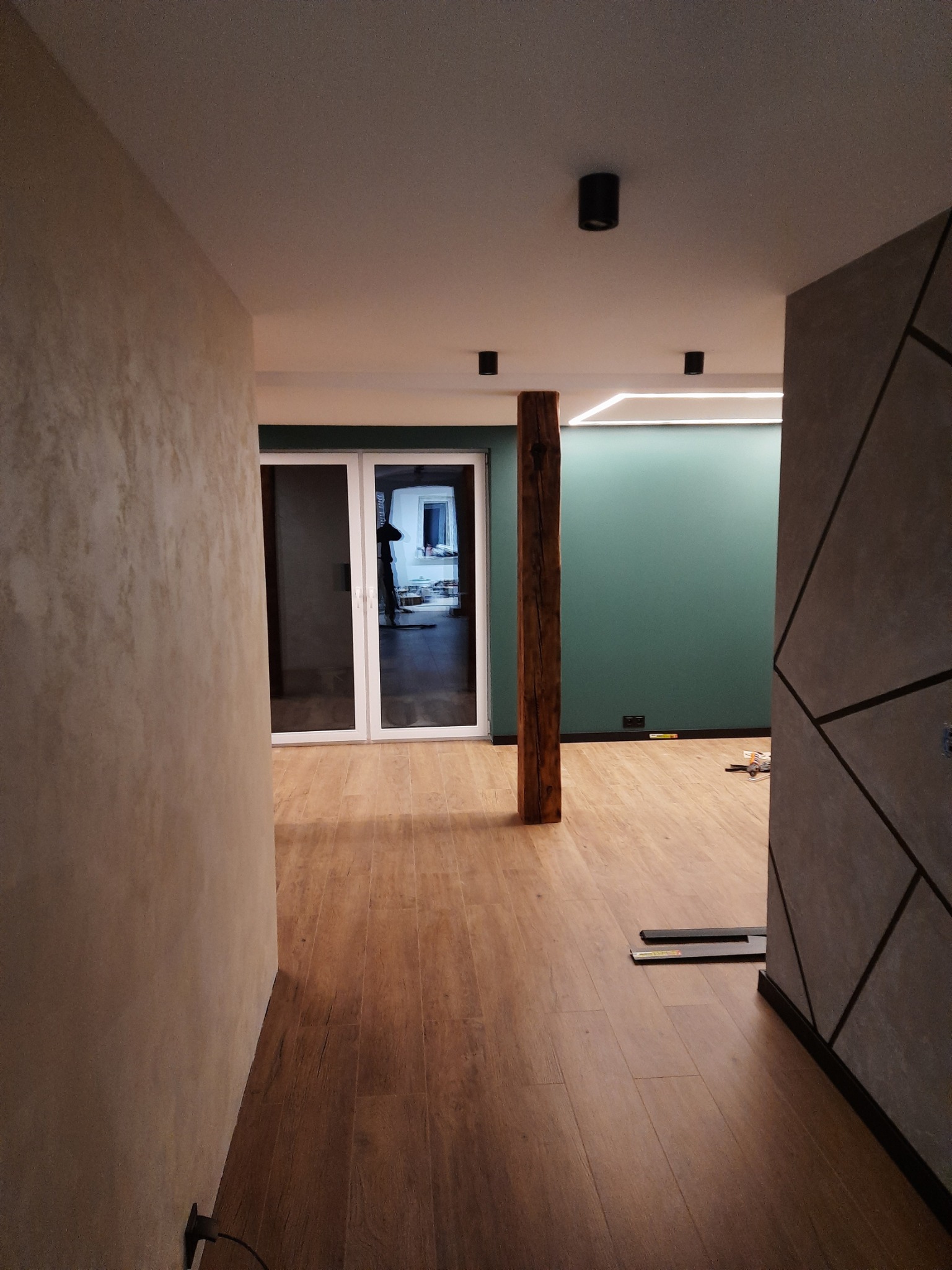 Wnętrze w trakcie wykańczania: widok na korytarz z drewnianą podłogą, ścianą z dekoracyjną fakturą i zieloną ścianą z oświetleniem LED, drewniany słup konstrukcyjny oraz narzędzia na podłodze.