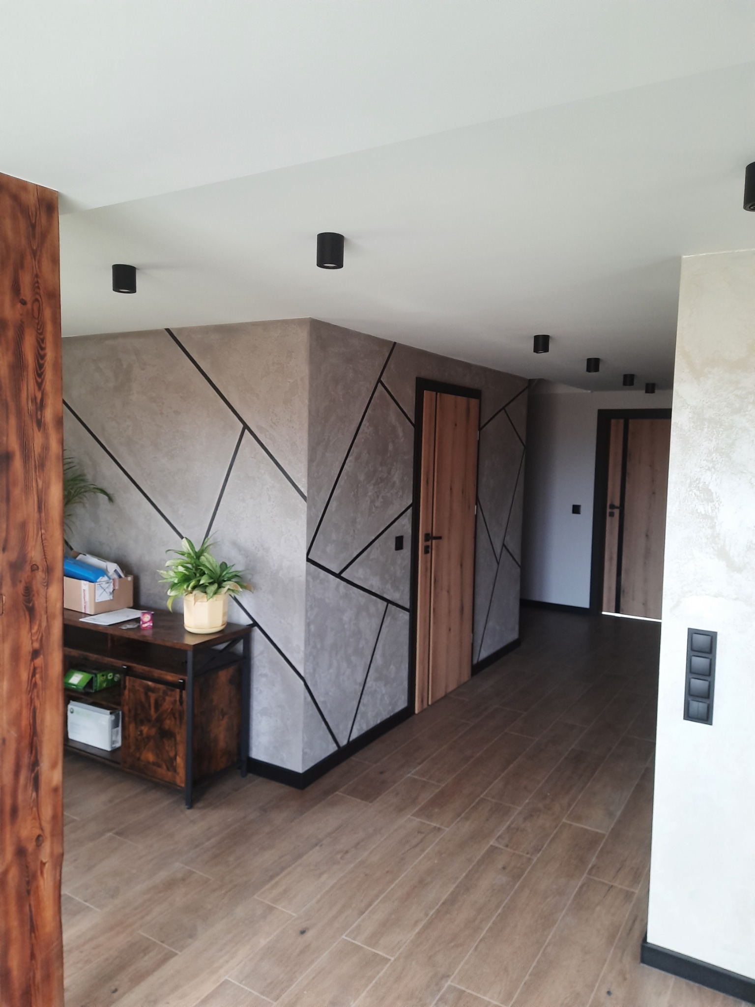 Nowoczesny korytarz z geometryczną dekoracją ścian w odcieniach szarości, drewnianą podłogą imitującą panele, czarnymi punktowymi lampami sufitowymi i drewnianymi drzwiami z czarnymi framugami.