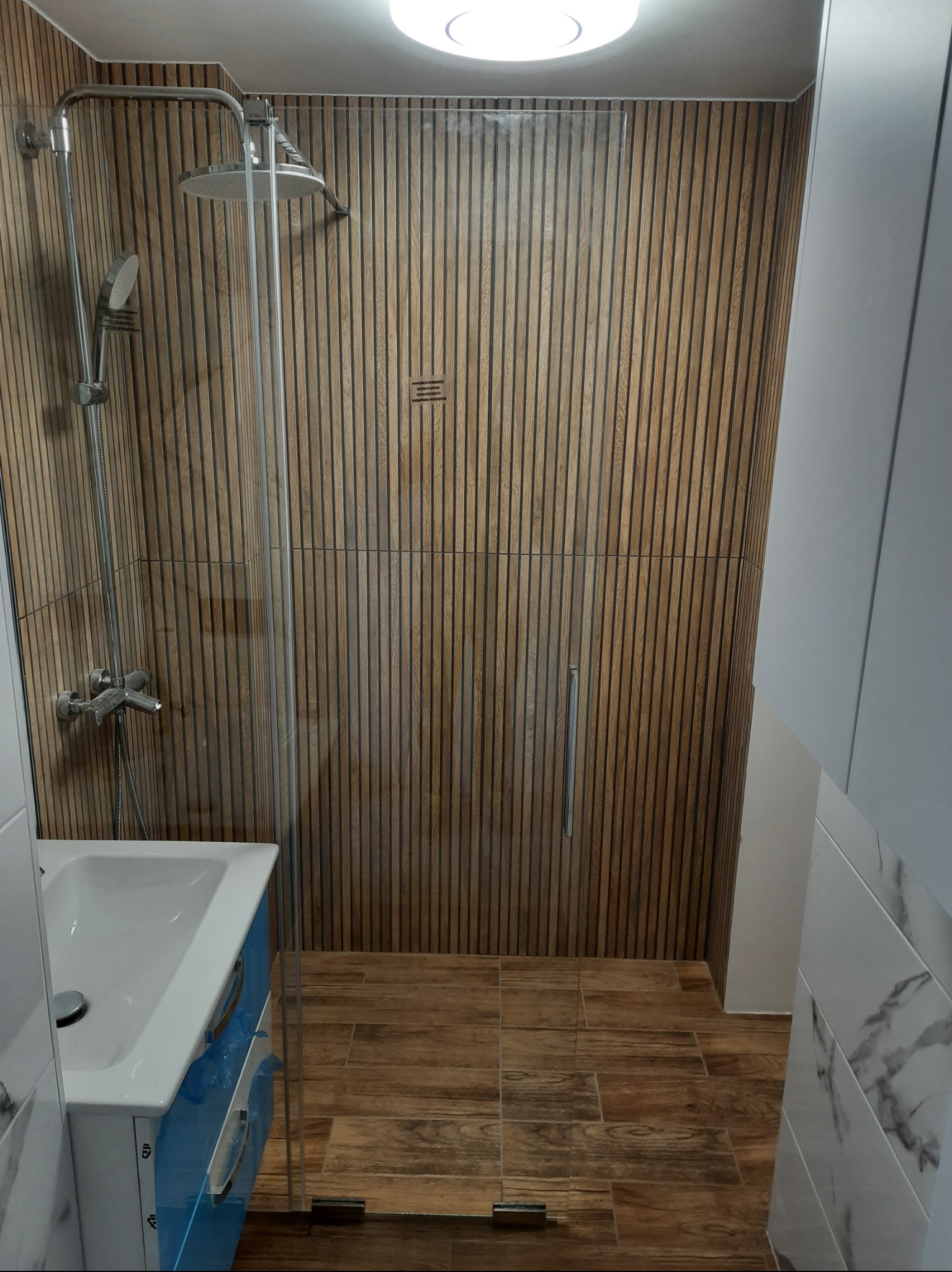 Nowoczesna kabina prysznicowa z drewnopodobnymi panelami ściennymi i podłogą, bateria prysznicowa z deszczownicą, umywalka z niebieską szafką. Całość w jasnym, minimalistycznym stylu.