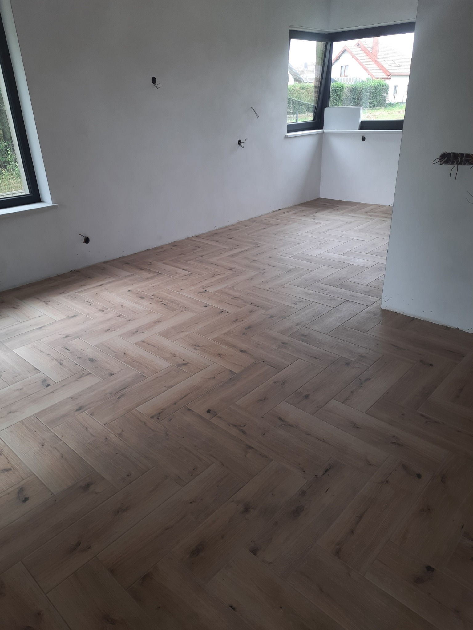 Ukończona podłoga z paneli ułożonych w jodełkę w jasnym, pustym pokoju z oknami i przygotowanymi instalacjami elektrycznymi na ścianach.