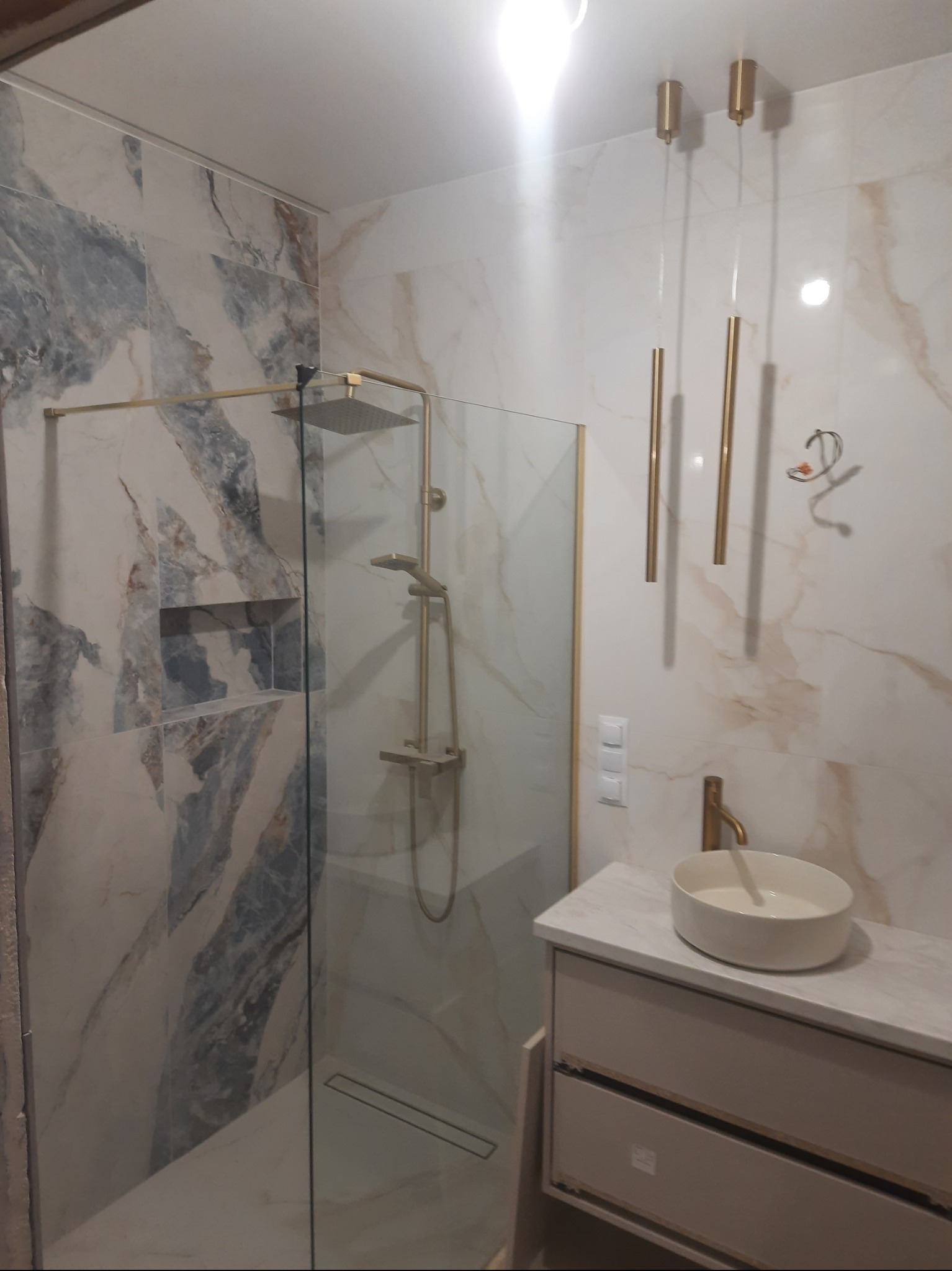 Wykończona łazienka z kabiną typu walk-in, marmurowymi płytkami w odcieniach bieli i szarości, złotą armaturą i wiszącymi lampami. Nowoczesny design.