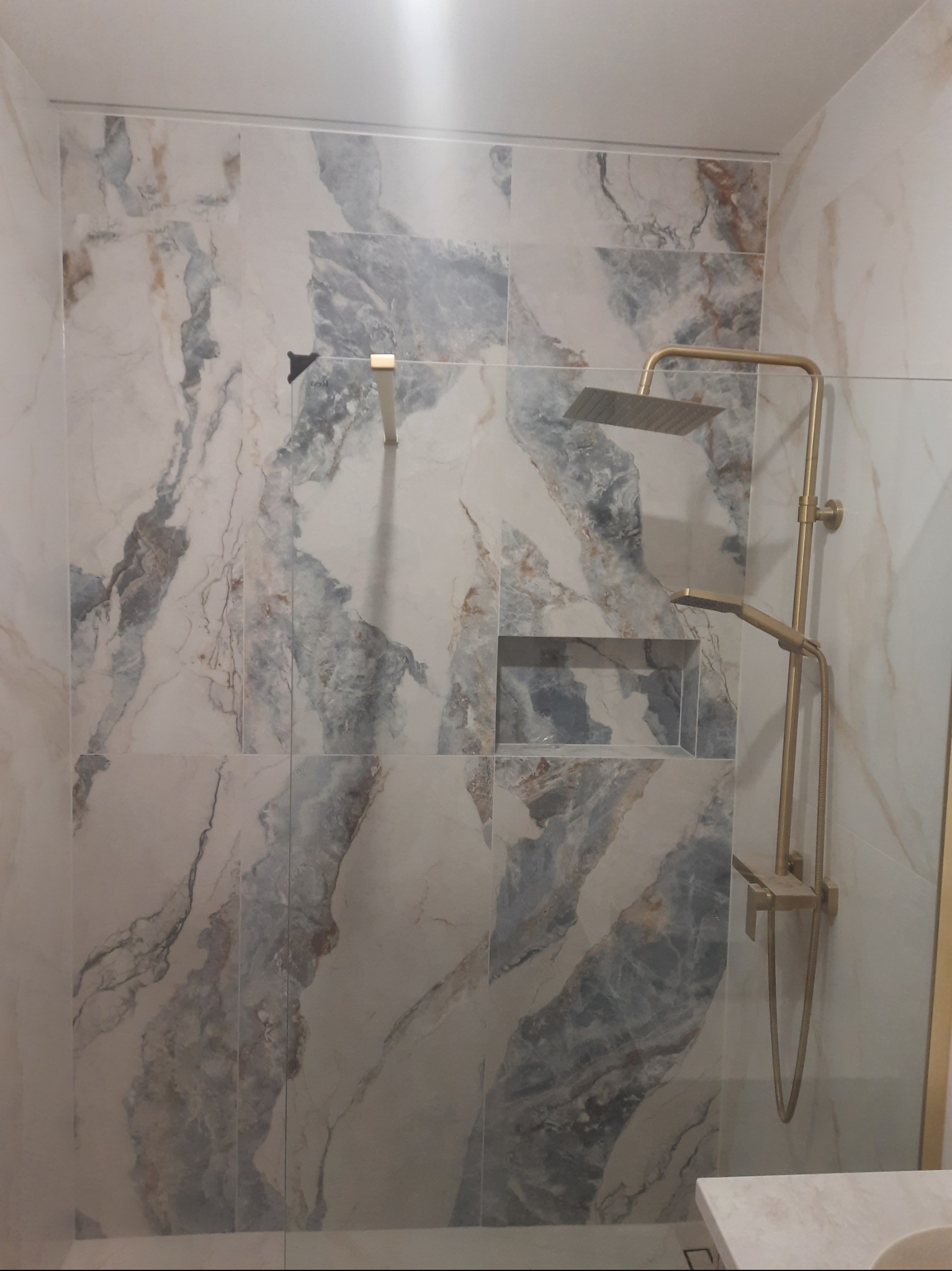 Wykończona łazienka z marmurowymi płytkami w odcieniach bieli i szarości, z wnęką i złotą armaturą prysznicową. Nowoczesny design.