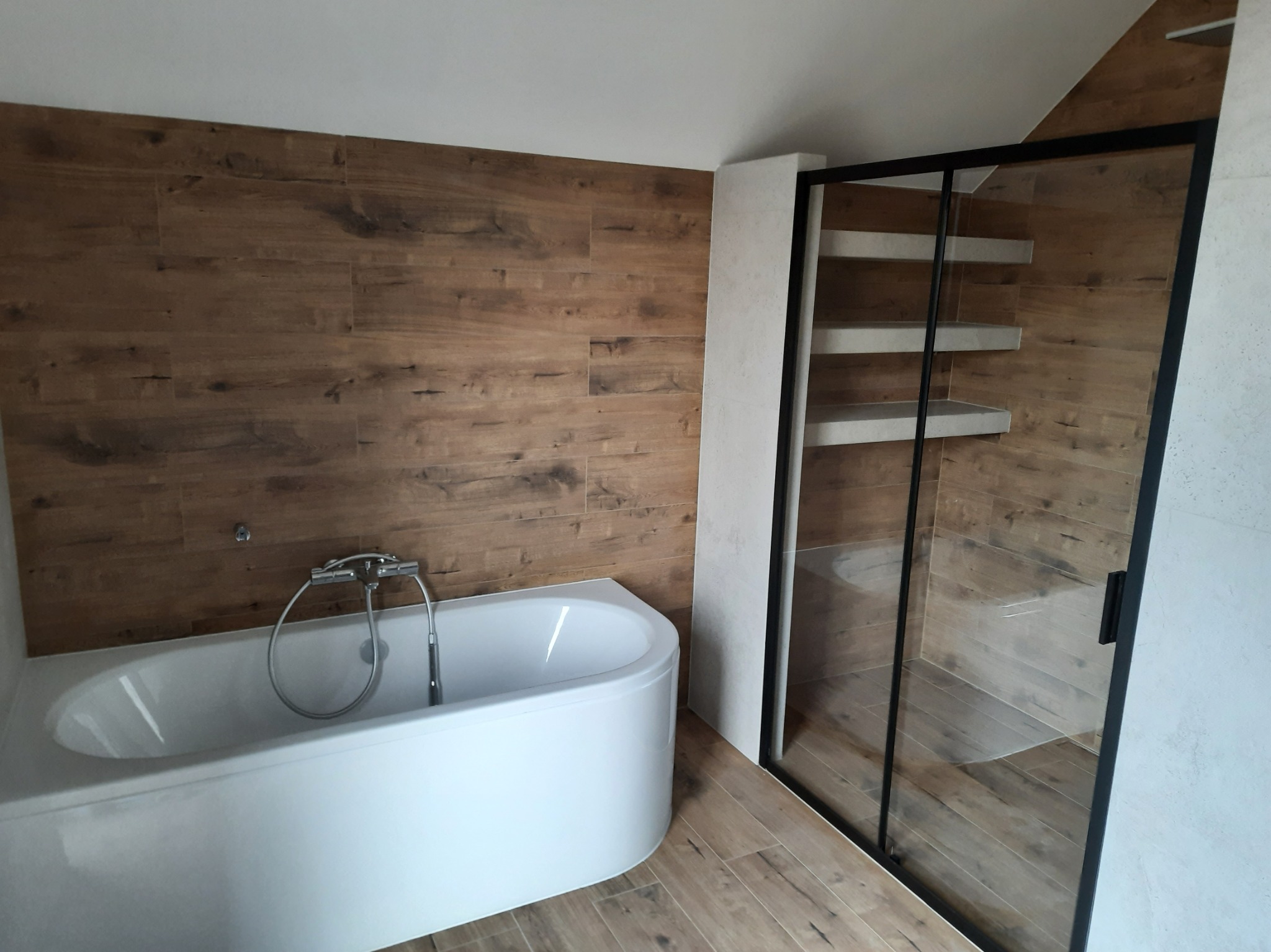 Nowoczesna łazienka z wanną i kabiną prysznicową typu walk-in, wykończona płytkami imitującymi drewno. Minimalistyczny design, czarne ramy kabiny, jasne ściany.