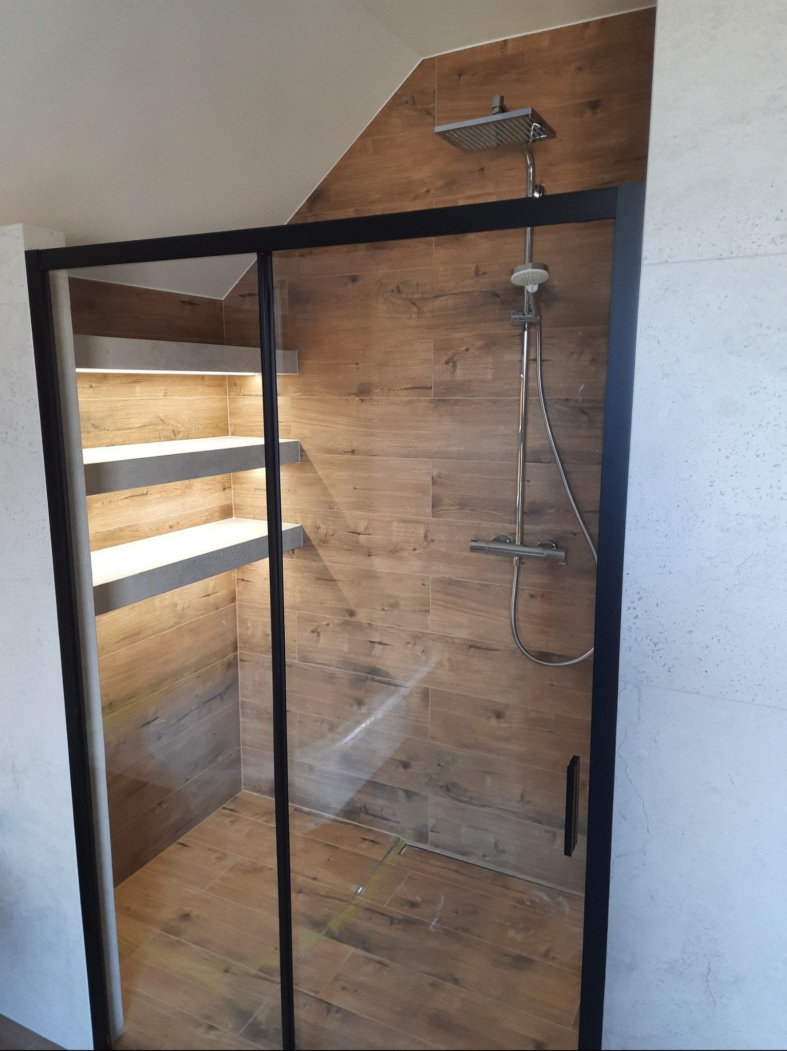 Nowoczesna kabina prysznicowa z czarną ramą, wykończona płytkami imitującymi drewno. Wewnątrz półki z podświetleniem LED. Minimalistyczny design, jasne oświetlenie.