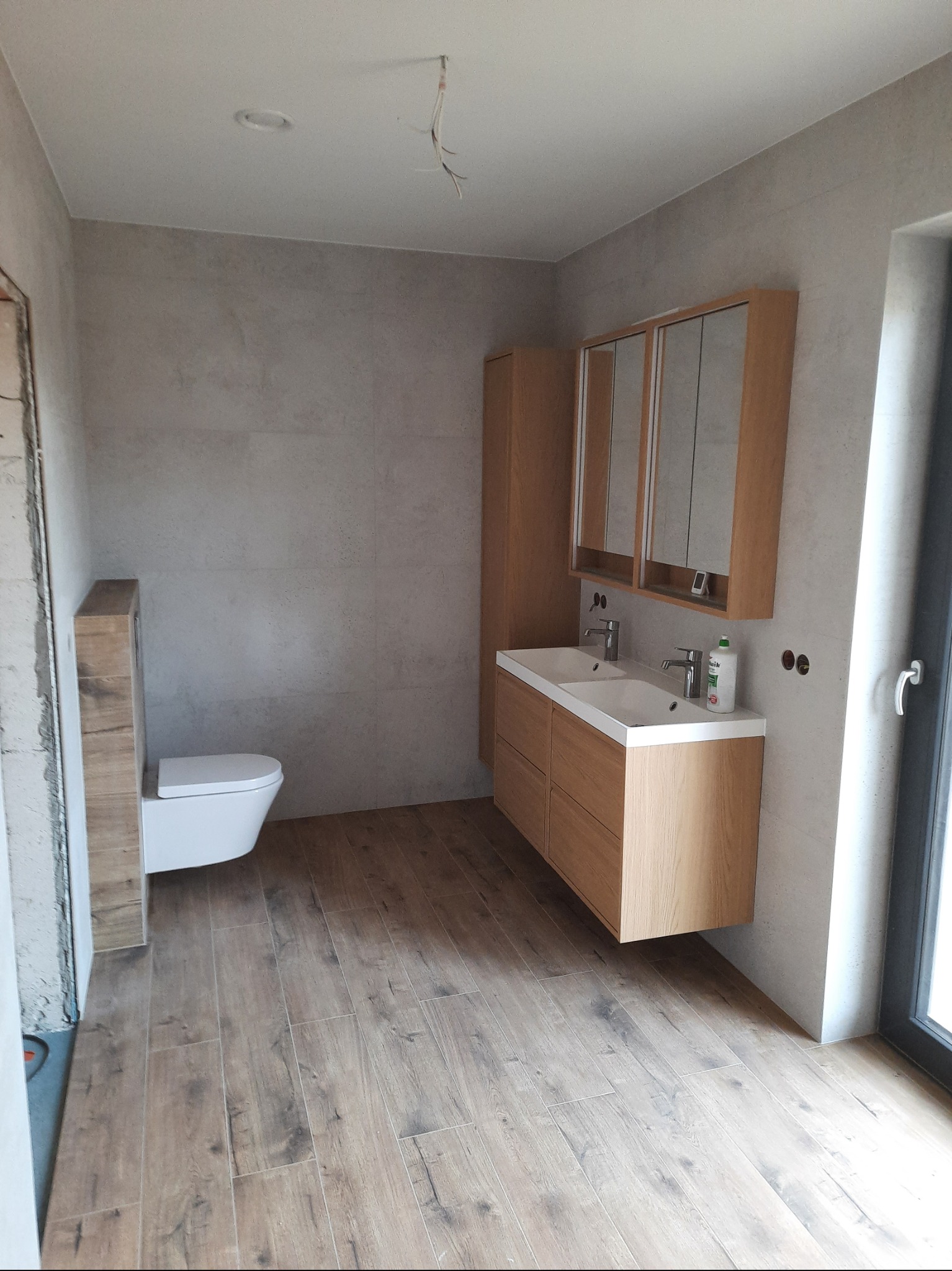 Jasna łazienka z drewnopodobną podłogą i szarą glazurą. Umywalka z szafką z jasnego drewna, lustro z szafkami. Widoczne niedokończone prace przy drzwiach.