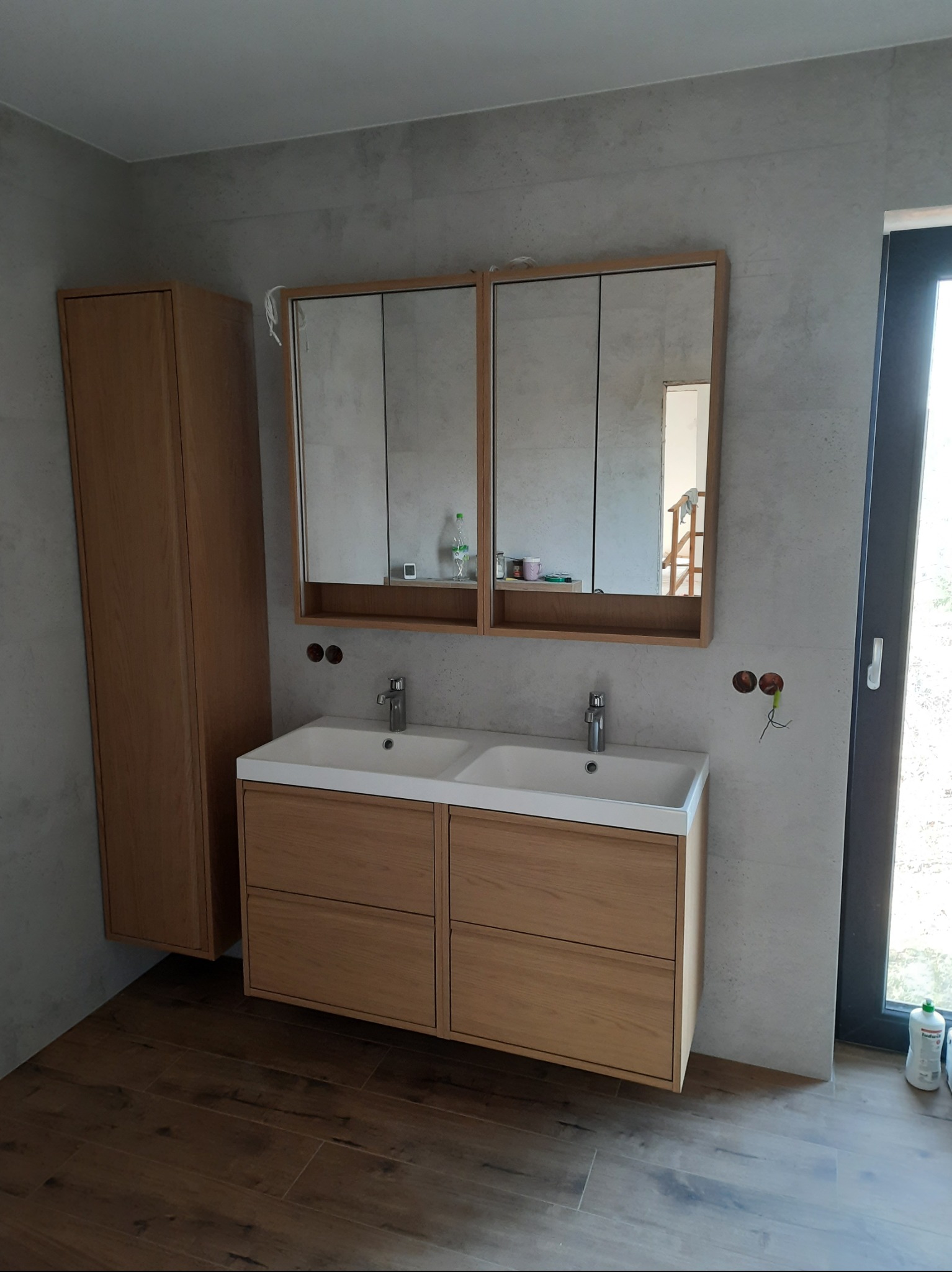 Nowoczesna łazienka z drewnianą szafką pod umywalką z dwiema misami, lustrem w drewnianej ramie i wysoką szafką. Szare płytki na ścianach i brązowe na podłodze.