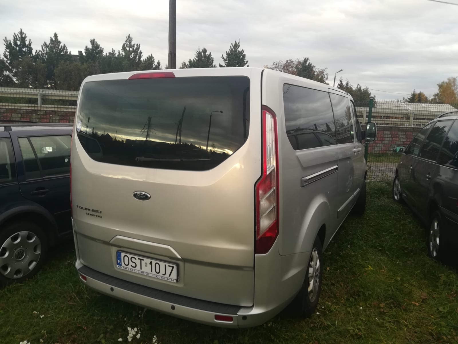 Srebrny van Ford Tourneo Custom zaparkowany na trawie, widok z tyłu, z widoczną tablicą rejestracyjną OST 10J7 i odbiciem krajobrazu w szybie.
