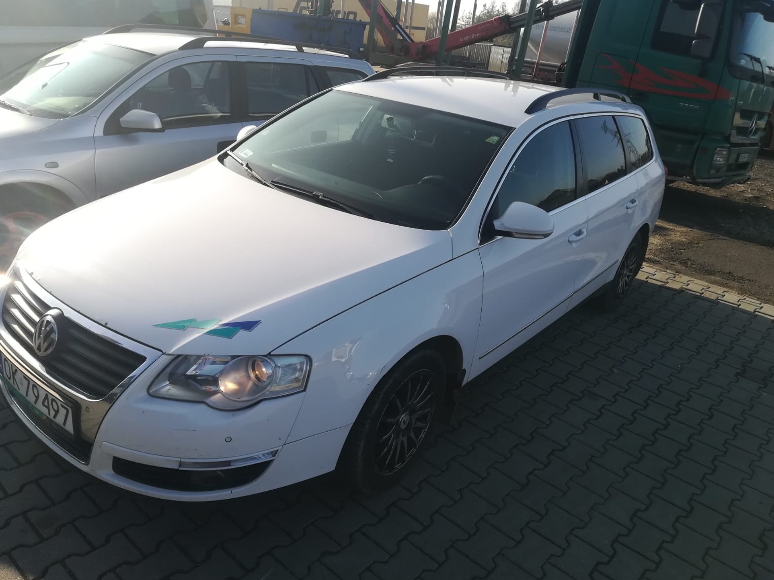 Biały samochód kombi z relingami dachowymi i naklejką z logo na masce, zaparkowany na kostce brukowej obok innego auta i ciężarówki.