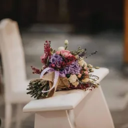 Bukiet ślubny z fioletowymi i różowymi kwiatami, przewiązany jutowym materiałem i fioletową wstążką, leży na białym krześle w kościele.